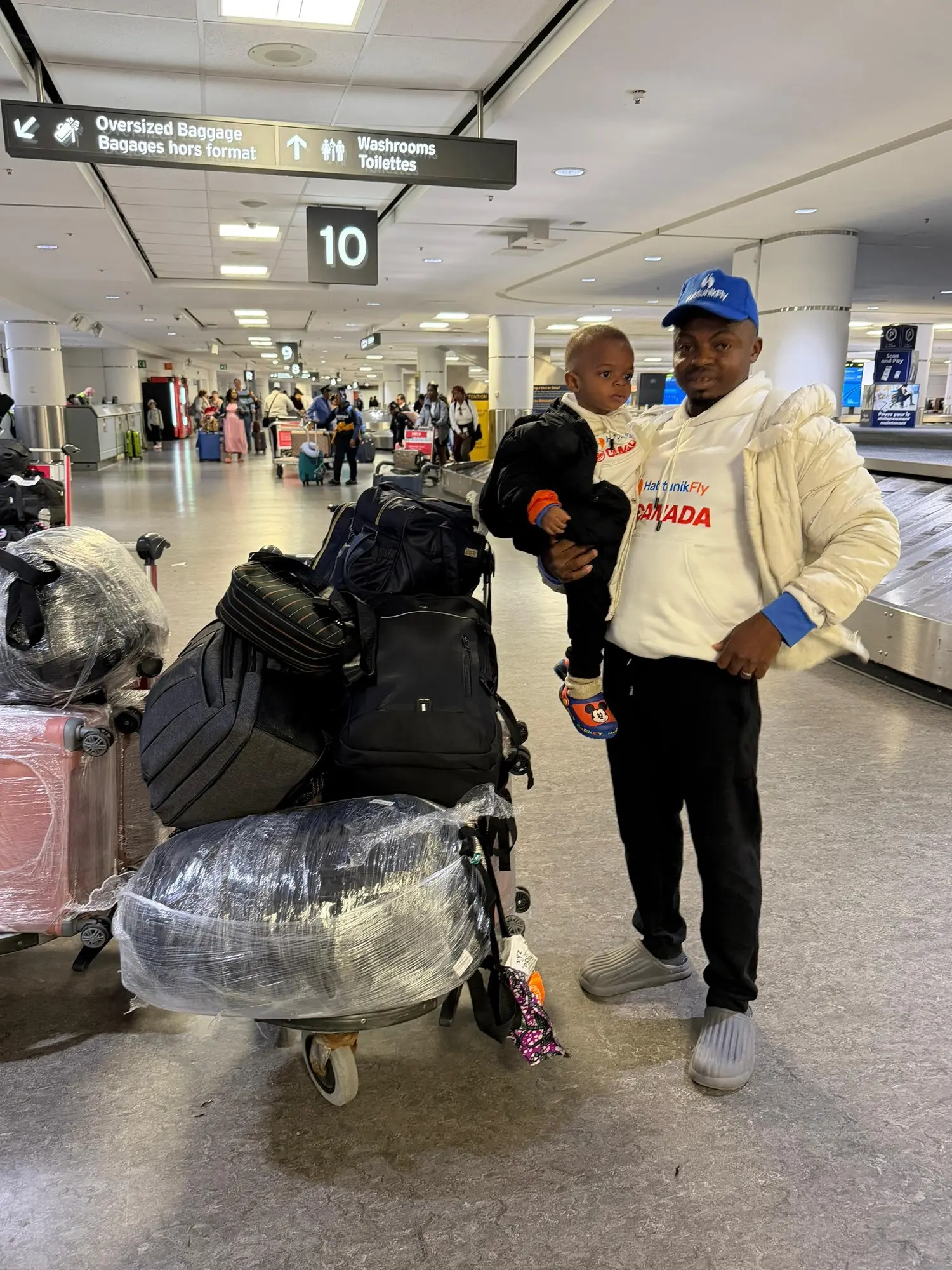 Client carrying his child beside a trolley of luggage in a Canadian airport arrivals hall